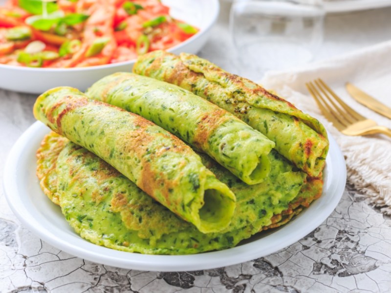 Mehrere gerollte Bärlauch-Pfannkuchen auf einem Stapel weiterer Pfannkuchen. Im Hintergrund ist ein Salat mit Tomaten zu sehen.