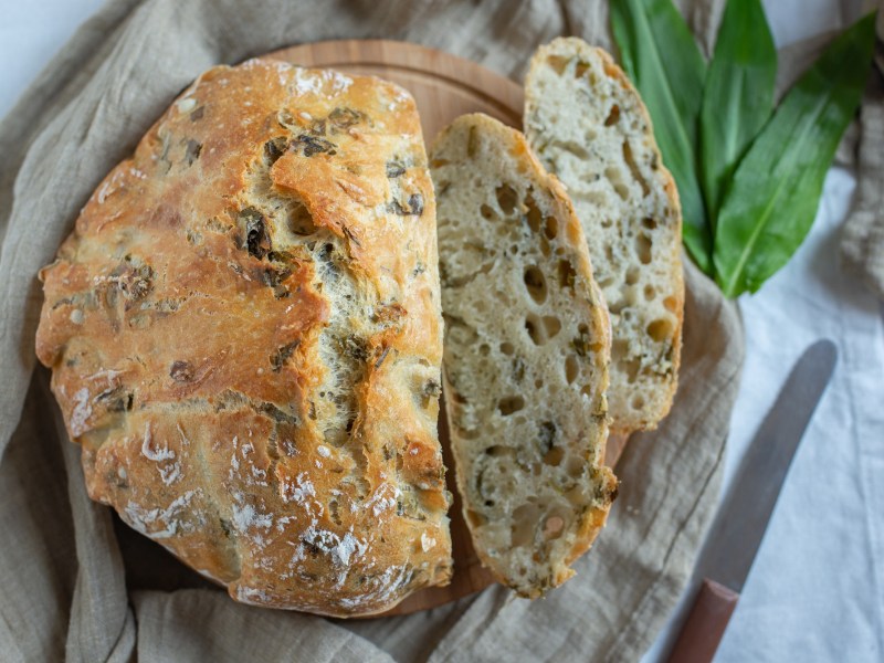 Ein angeschnittenes rundes Bärlauchbrot auf einem Brett, zwei Scheiben und ein Messer sowie Bärlauch daneben. Draufsicht.