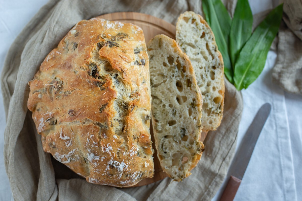 Ein angeschnittenes rundes Bärlauchbrot auf einem Brett, zwei Scheiben und ein Messer sowie Bärlauch daneben. Draufsicht.