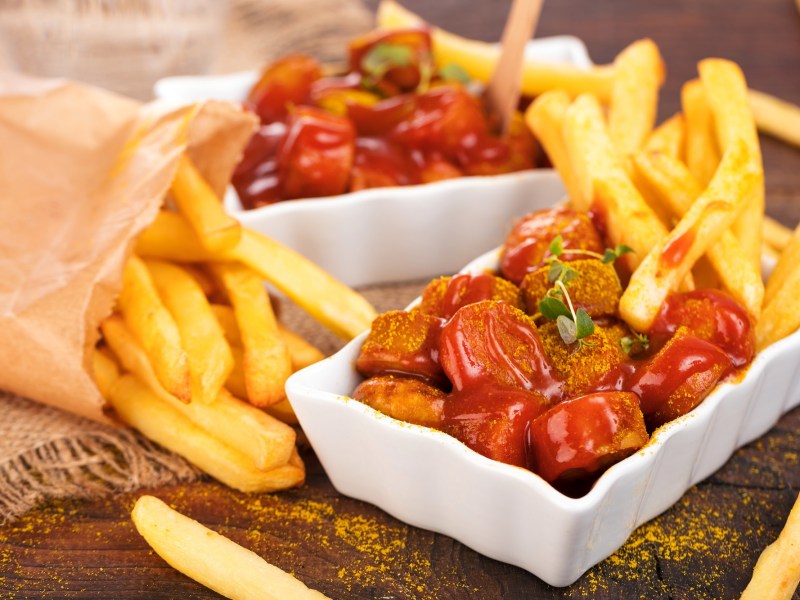 Berliner Currywurst mit Pommes in einer weißen Porzellanschale. Daneben eine Tüte Pommes.