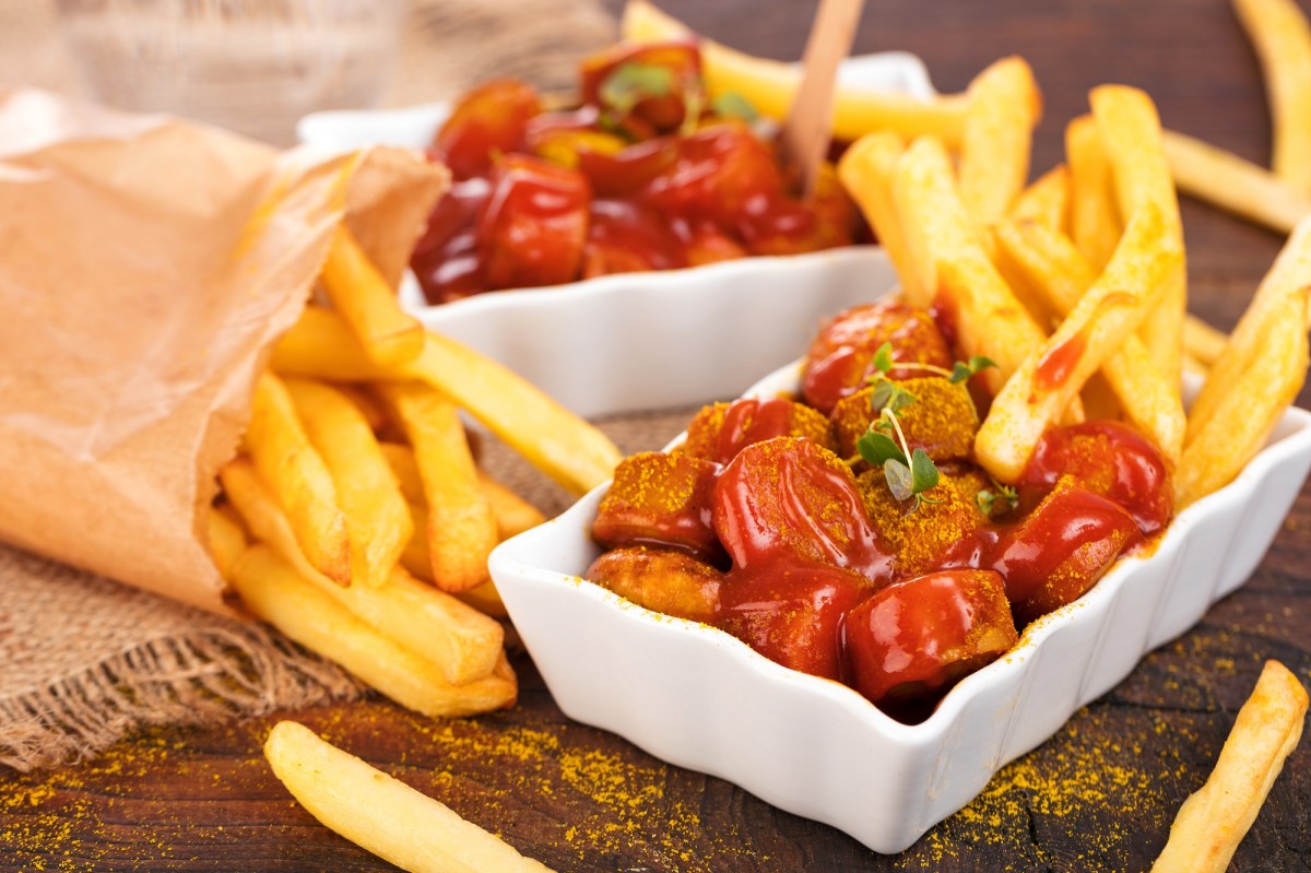 Berliner Currywurst mit Pommes in einer weißen Porzellanschale. Daneben eine Tüte Pommes.