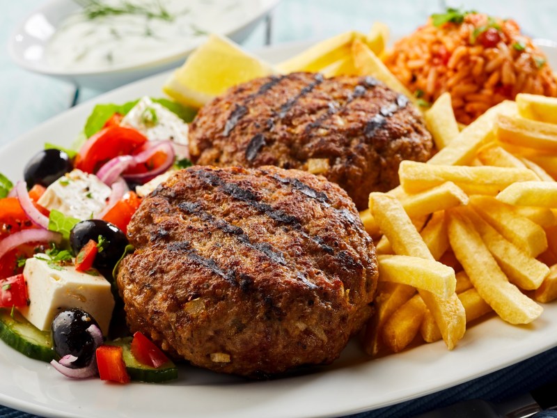 Bifteki auf einem Teller mit Pommes, Tomatenreis und Tomaten-Schafskäse-Salat.