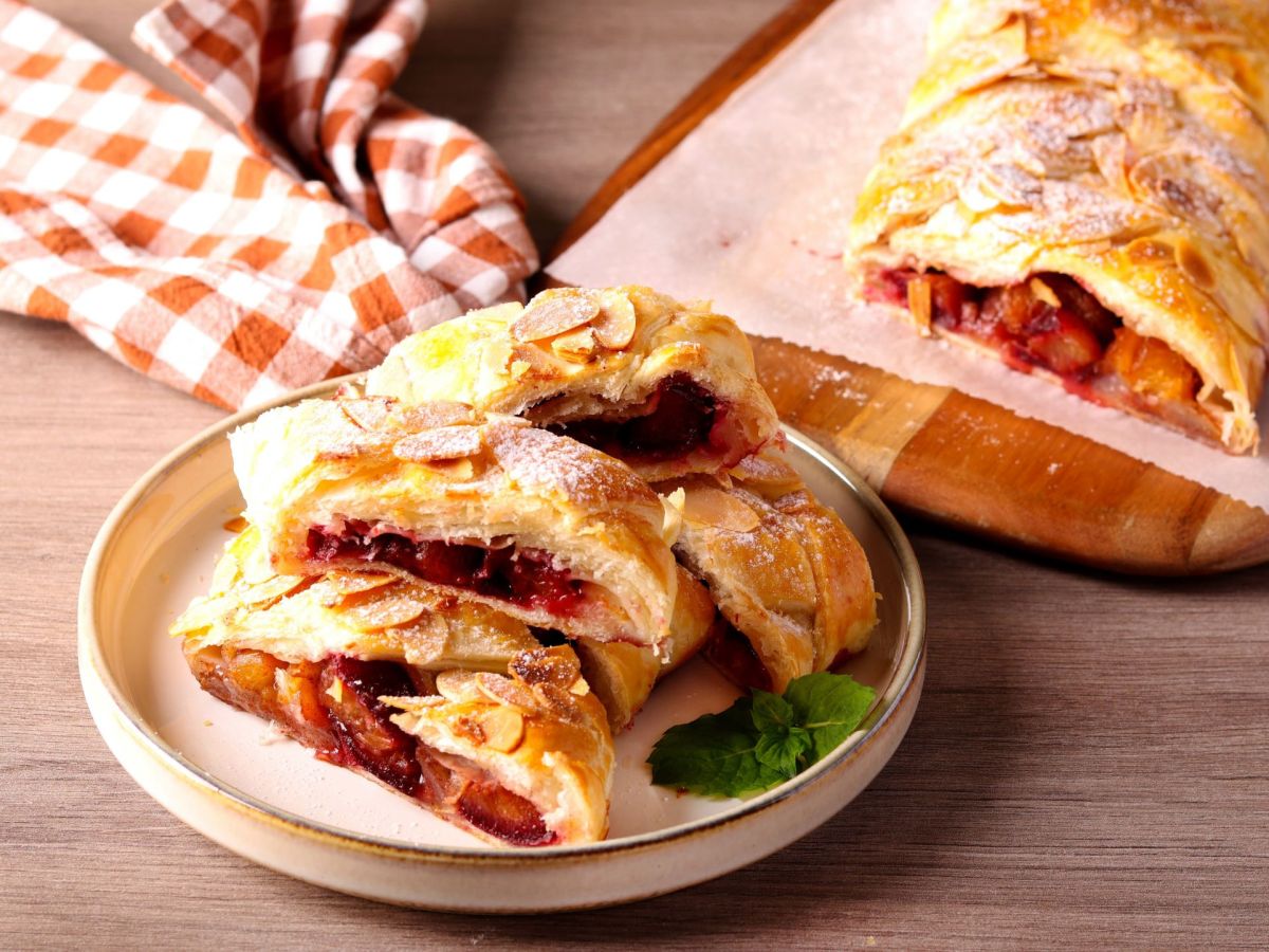 Blätterteigzopf mit Pfirsich, aufgeschnitten auf einem Teller. Im Hintergrund ist der restliche Strudel zu sehen.