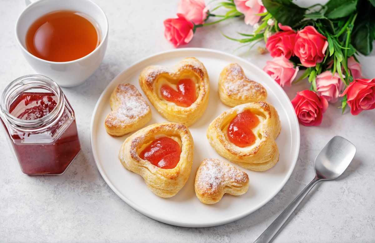 Teller mit Blätterteig-Herzen mit Marmelade, Tee und Rosen auf hellem Untergrund.