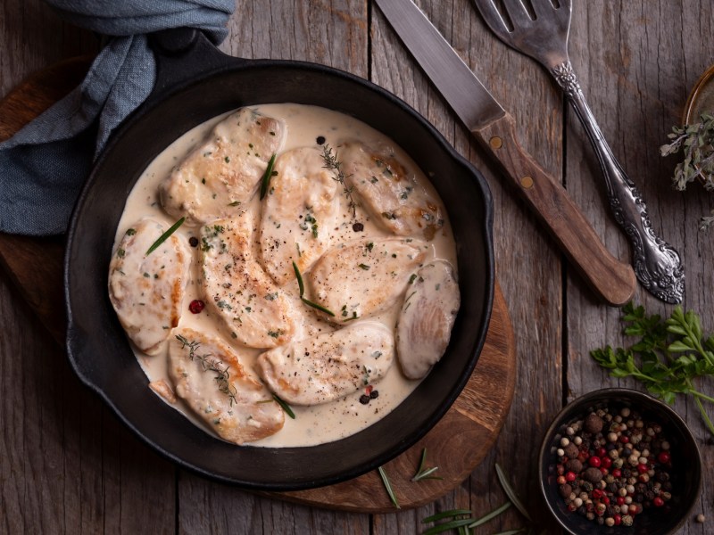 Chicken Supreme in einer gusseisernen Pfanne auf einem Holzbrett. Daneben liegt ein Messer und eine Schale mit Pfefferkörnern.