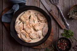 Chicken Supreme in einer gusseisernen Pfanne auf einem Holzbrett. Daneben liegt ein Messer und eine Schale mit Pfefferkörnern.