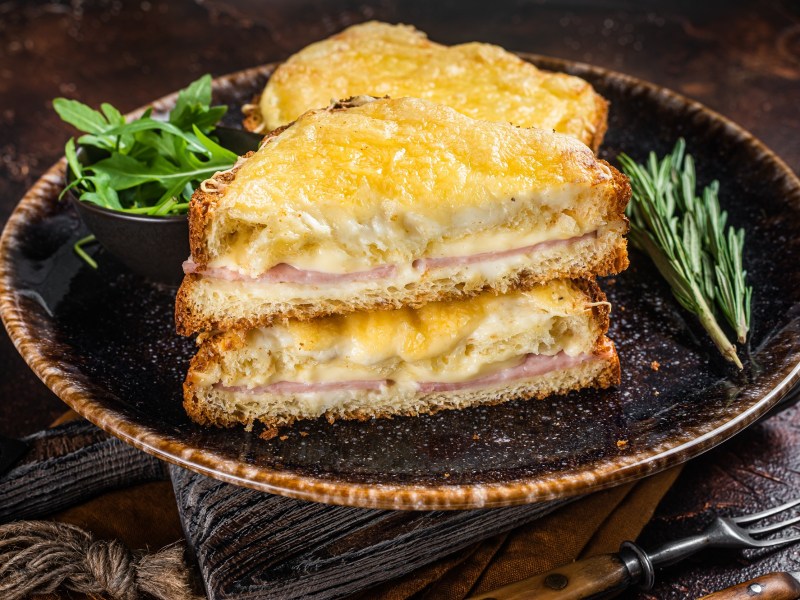 Zwei diagonal durchgeschnittene Croque Monsieur auf einem dunklen Teller. Daneben liegen ein paar Zweige Rosmarin sowie eine Schale Rucola.