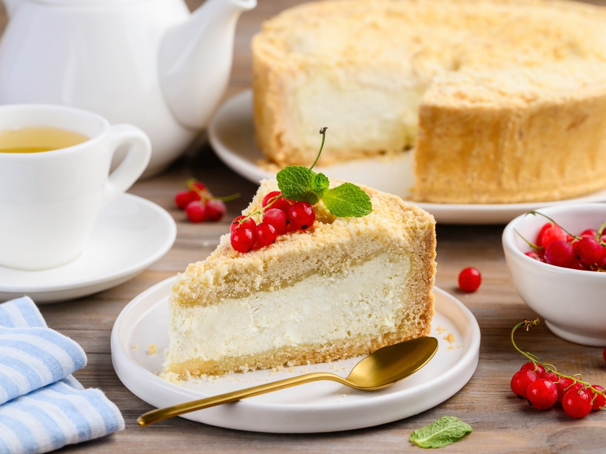 Ein Stück Dreh-dich-um-Kuchen, garniert mit Johannisbeeren und Minze, auf einem weißen Teller, Daneben liegt ein goldener Löffel. Im Hintergrund unscharf der Rest des Kuchens.