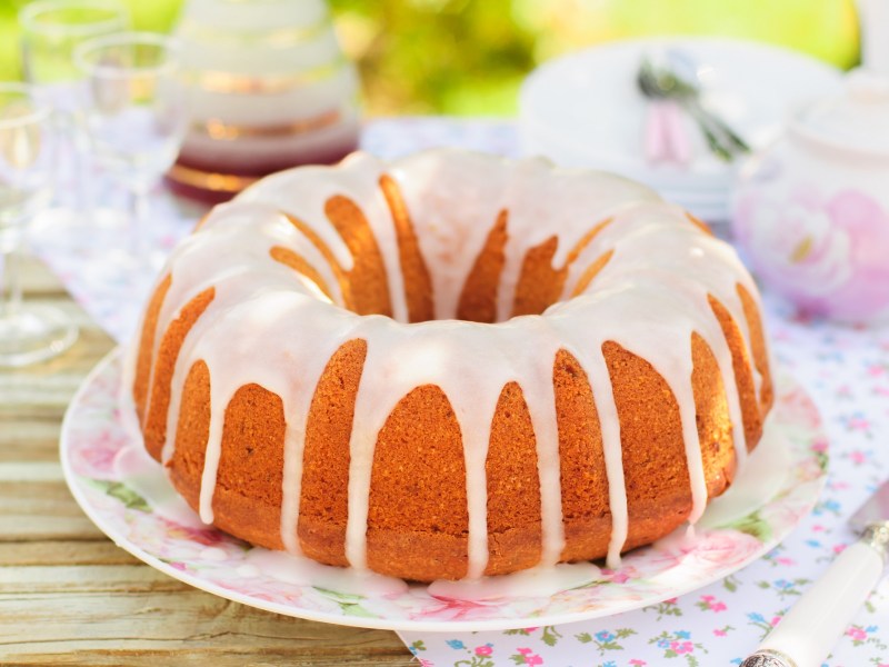 Ein Eierlikörkuchen mit Guss auf einem Blumenteller. Drum herum sind Blumenservietten und Sonnenlicht fällt auf den Kuchen.