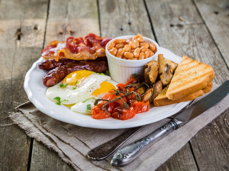 English Breakfast mit Bacon, Würtchen, Spiegeleiern, Baked Beans, Tomaten, Pilzen ud Toast auf einem weißen Teller. Daneben Messer und Gabel.