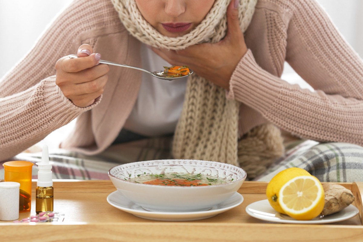 Eine Frau mit Schal isst eine Suppe, ein gutes Essen bei Erkältung. Neben ihr liegen Ingwer, Zitronen und Medikamente.