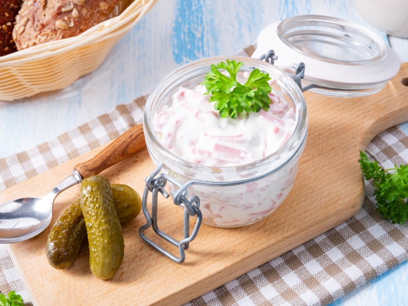 Ein Bügelglas mit Fleischsalat und Gewürzgurken daneben auf einem Holzbrett.