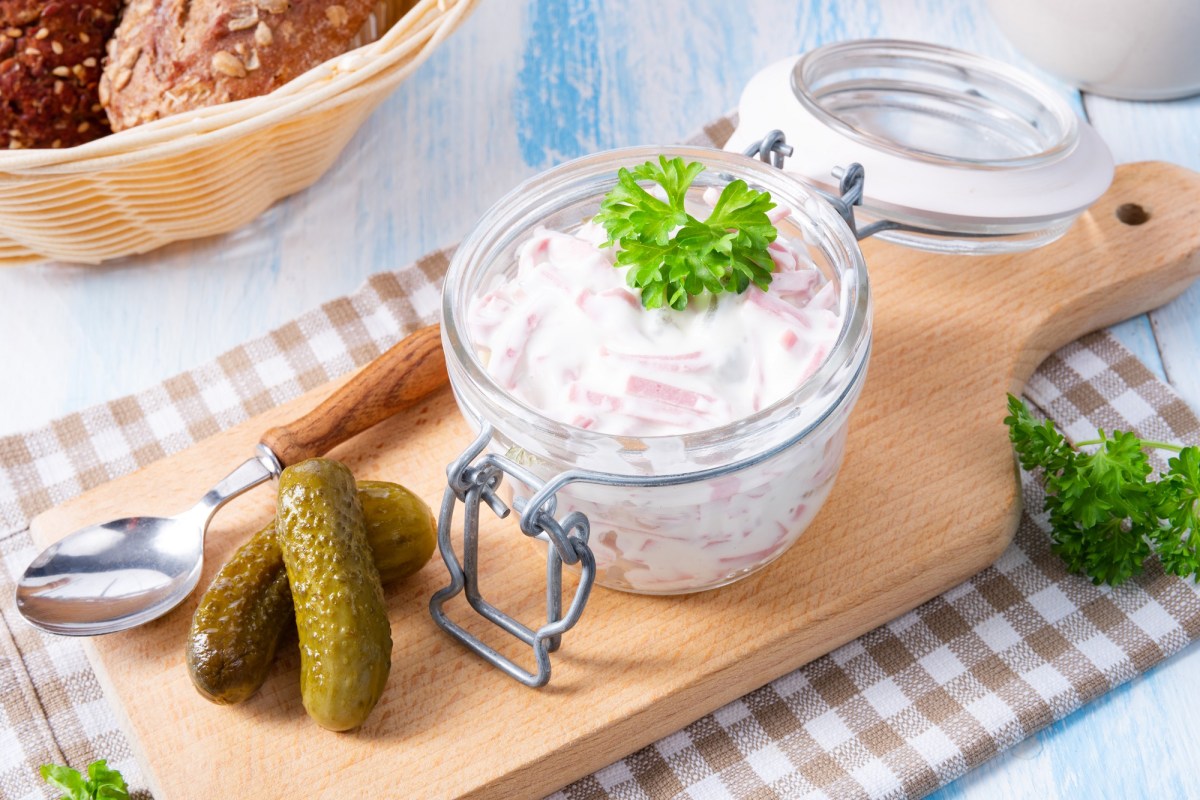 Ein Bügelglas mit Fleischsalat und Gewürzgurken daneben auf einem Holzbrett.