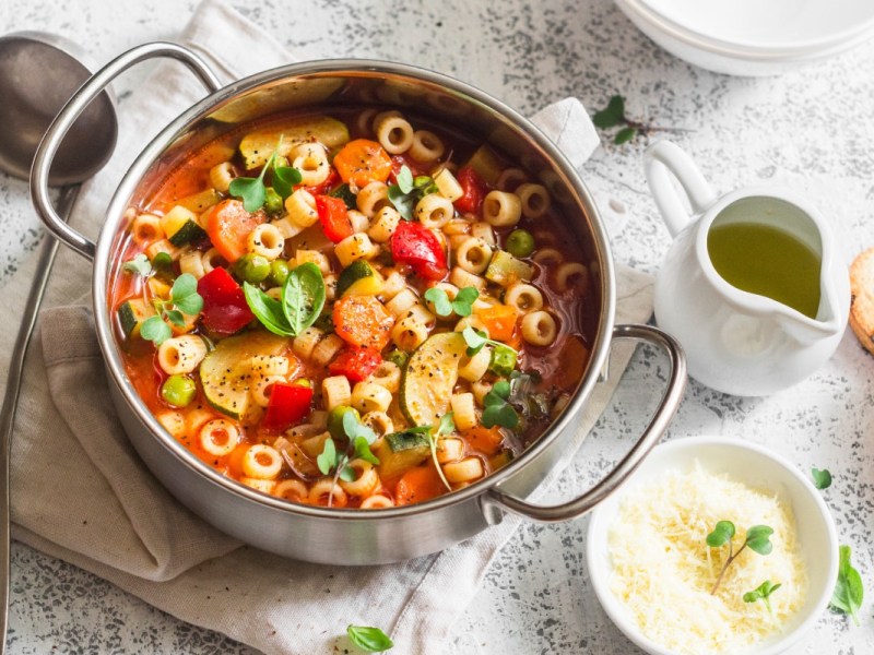 Ein Topf mit Frühlings-Minestrone in der Draufsicht. Daneben liegt eine Kelle und es stehen ein Schälchen mit Käse und eine Kanne mit Öl herum.