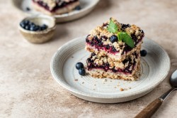 3 Frühstücksriegel mit Blaubeeren auf einen Teller gestapelt, dekoriert mit einem Minzblättchen, daneben eine Gabel., dahinter ein weiterer Teller.