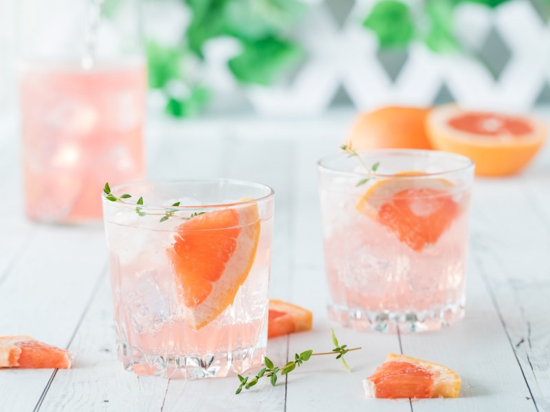 Drei Gläser Grapefruit Fizz mit halben Grapefruits auf hellem Untergrund.