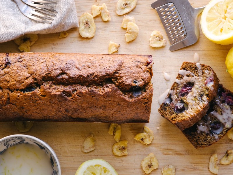 Ein Himbeer-Bananenbrot, von dem ein Stück abgeschnitten wurde in der Draufsicht. Drumherum liegen Küchenwerkzeuge und Zutaten.