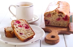 Ein angeschnittener Himbeerkuchen mit weißer Schokolade auf einem Brett, daneben ein Teller mit zwei Stücken Kuchen und eine Tasse.