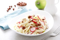 Eine weiße Schale mit Kohlrabi-Apfel-Salat, Nüsse und Apfel auf hellem Tisch