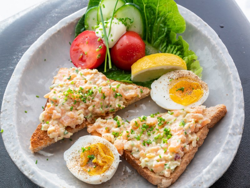 Ein Teller mit zwei Toastscheiben mit Lachs-aufstrich, Tomaten und zwei halben Eiern.