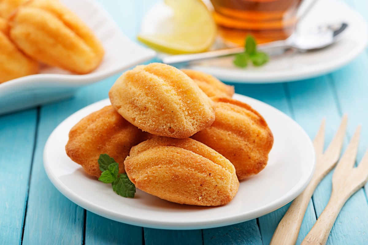 Madeleines backen: einige Madeleines liegen übereinander geschichten auf einem weißen Teller. Sie sind mit etwas Minze garniert. Dahinter sieht man eine Tasse Tee und noch mehr Madeleines. Nahaufnahme.