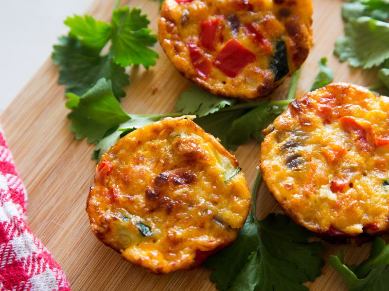 3 Omelett-Muffins mit Gemüse auf einem Brett, daneben Petersilie und ein rot-weiß kariertes Geschirrtuch, Draufsicht.