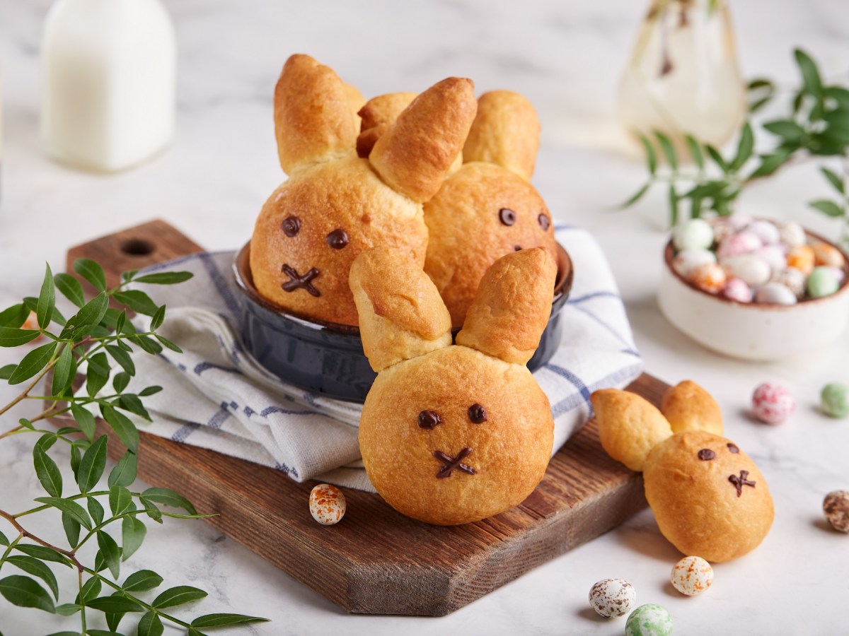 ein paar Osterhasen-Brötchen auf einem Brett miteinem Geschirrtuch, drumherum Osterdeko und Zweige.