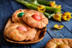 3 Osterkränze aus Hefeteig mit je einem bunten Ei in der Mitte auf einem Holzteller, daneben Osterglocken.