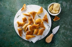 Pikante Blätterteig-Happen auf einem weißen Teller, der auf dunkelgrüner Unterlage steht. Daneben eine Schale mit Parmesan und ein Parmesan-Messer.