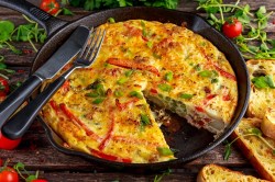 Quiche-Auflauf, von dem ein Stück fehlt, in einer gusseisernen Pfanne. Darauf liegen Messer und Gabel und drumherum ist der Tisch mit Brot und Rucola bedeckt.