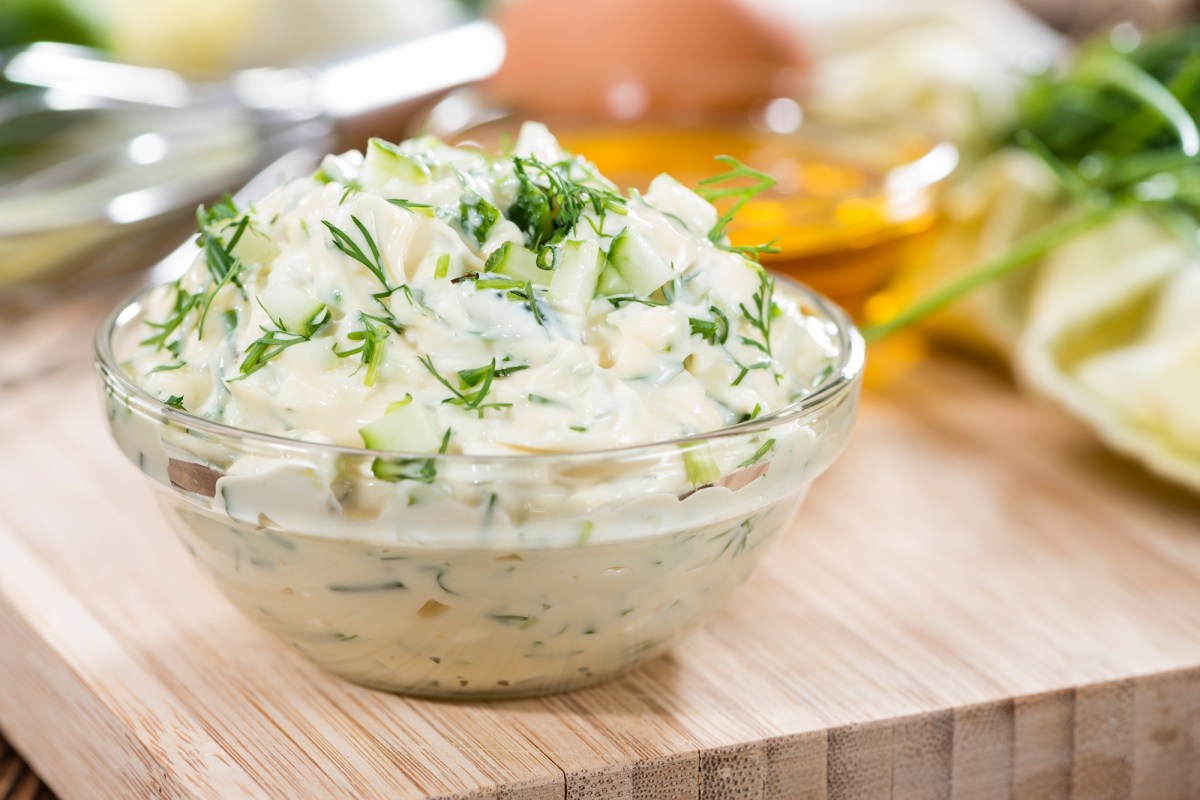 Remoulade in einer Glasschale, die auf einem Holzbrett steht. Dahinter sind unscharf die Zutaten zu erkennen.