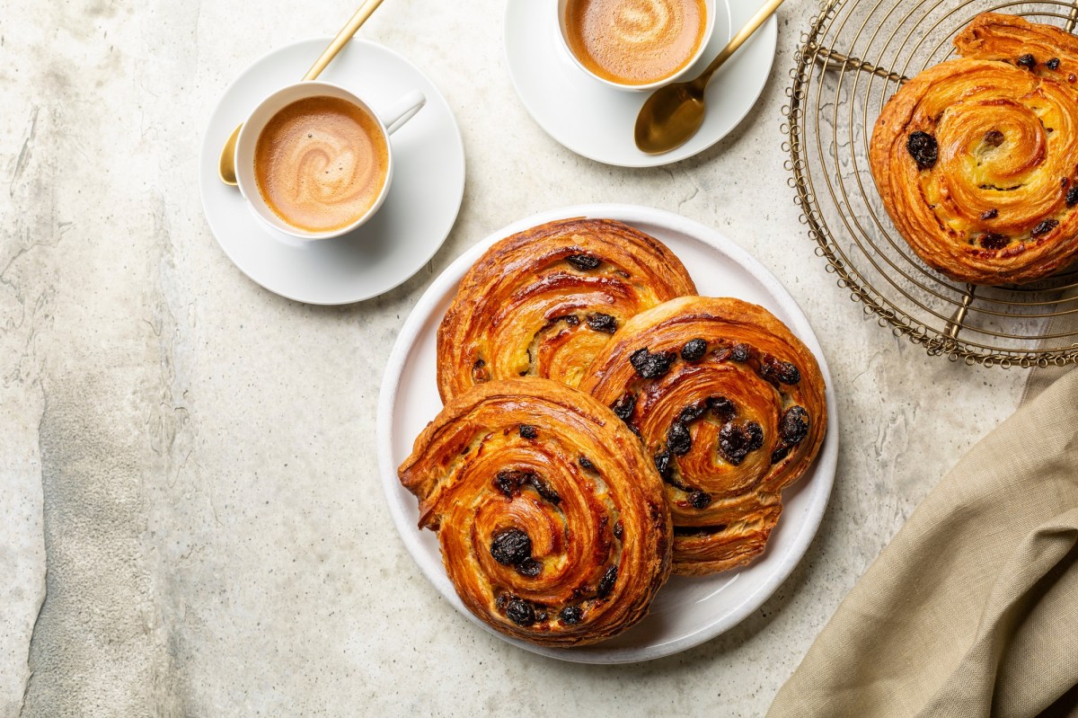 Rosinenschnecken auf einem weißen Teller. Dieser steht auf einem beigen Tischtuch. Daneben eine Tasse Kaffee und ein Gitter mit noch mehr Rosinenschnecken.