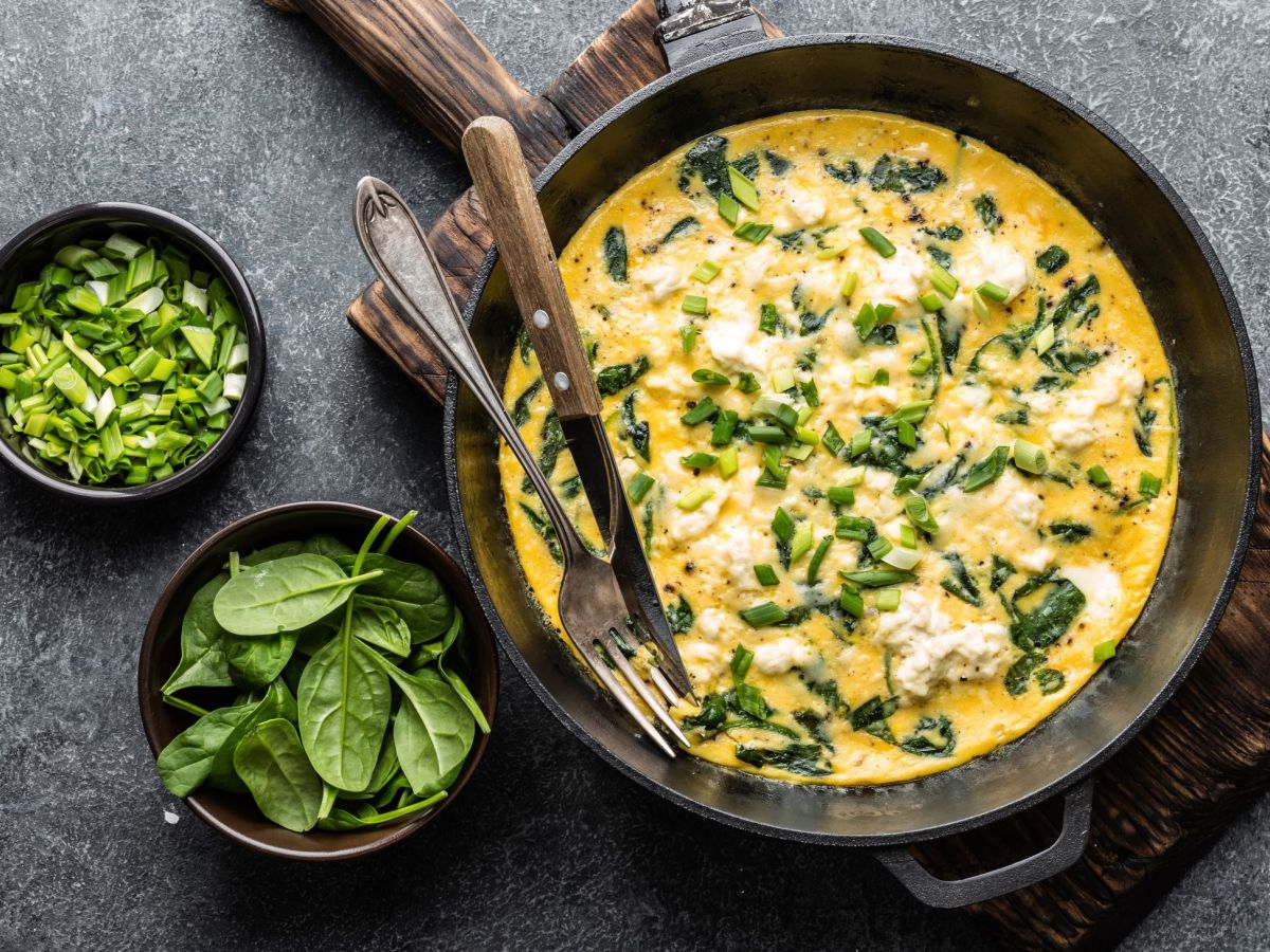 Rührei mit Feta und Spinat, angerichtet in einer großen Pfanne. Daneben stehen Frühlingszwiebeln und Spinat in zwei kleineren Schüsseln.