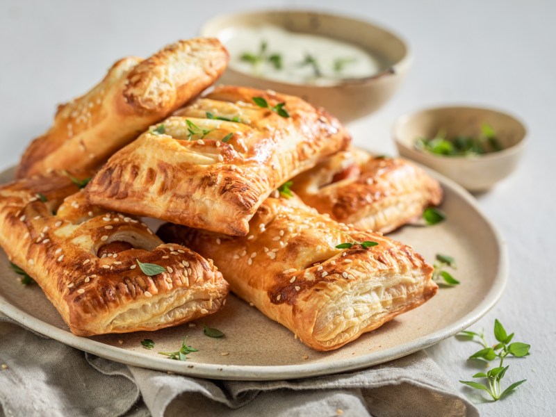 mehrere Spinat-Blätterteig-Taschen mit Feta auf einem Teller, im Hintergrund eine Schale mit Dip und eine mit Kräutern.