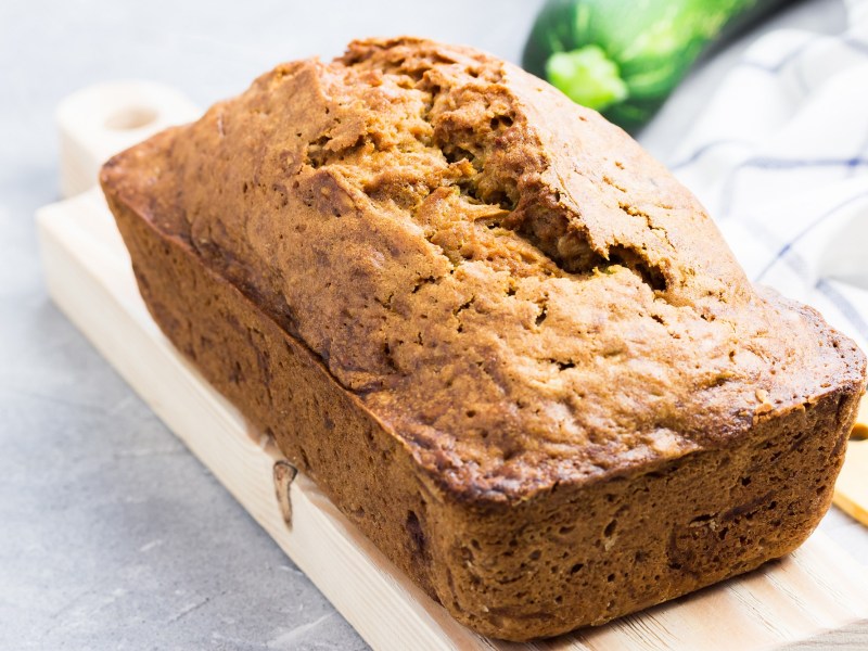 Süßes Zucchinibrot auf einem weißen Brett, im Hintergrund eine Zucchini.