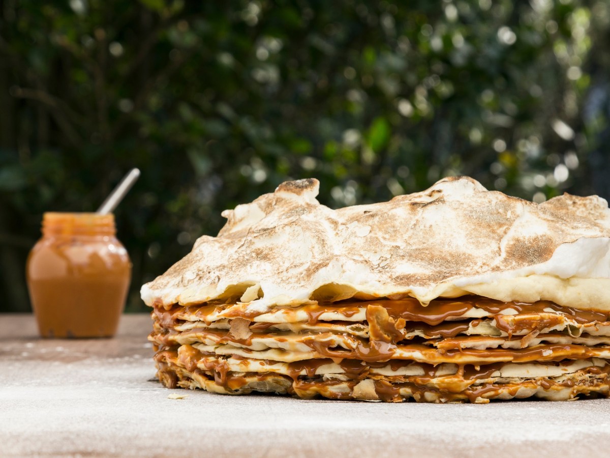 Tausend-Blatt-Torte mit Blätterteig und Baoserhaube auf einem Tisch. Daneben unscharf im Hintergrund ein Glas Dulce de Leche.