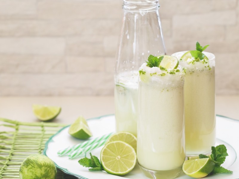 2 Gläser brasilianische Limonade neben einer Flasche auf einem runden Tablett. Daneben liegen Limettenscheiben und Minzblätter.