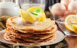 Ein Stapel britische Pancakes mit Zitronenstücken obendrauf. Im Hintergrund stehen die Zutaten.