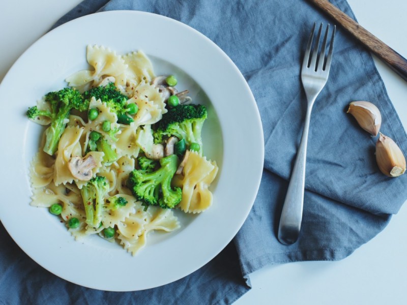 Ein Teller mit Brokkoli-Champignon-Pasta mit Erbsen. Daneben liegt Besteck.