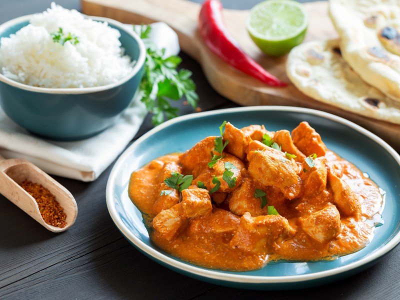 Butter Chicken auf einem hellblauen Teller. Im Hintergrund unscharf Reis und Naan.