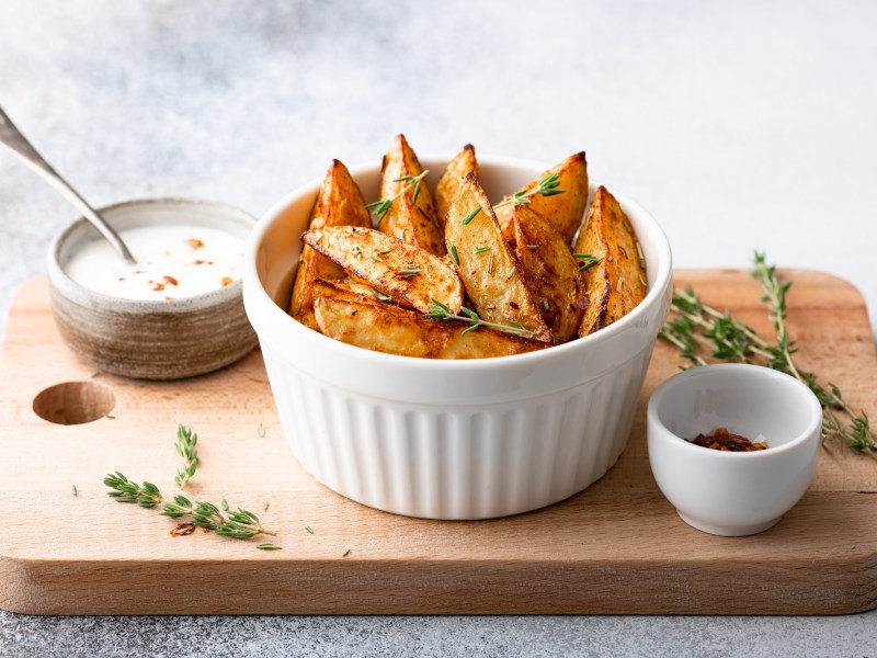 Eine weiße Schale mit Chilikartoffeln, in Wedges geschnitten, auf einem Brett, auf dem auch eine Schale Chiliflocken und eine Schale Dip mit Löffel steht.