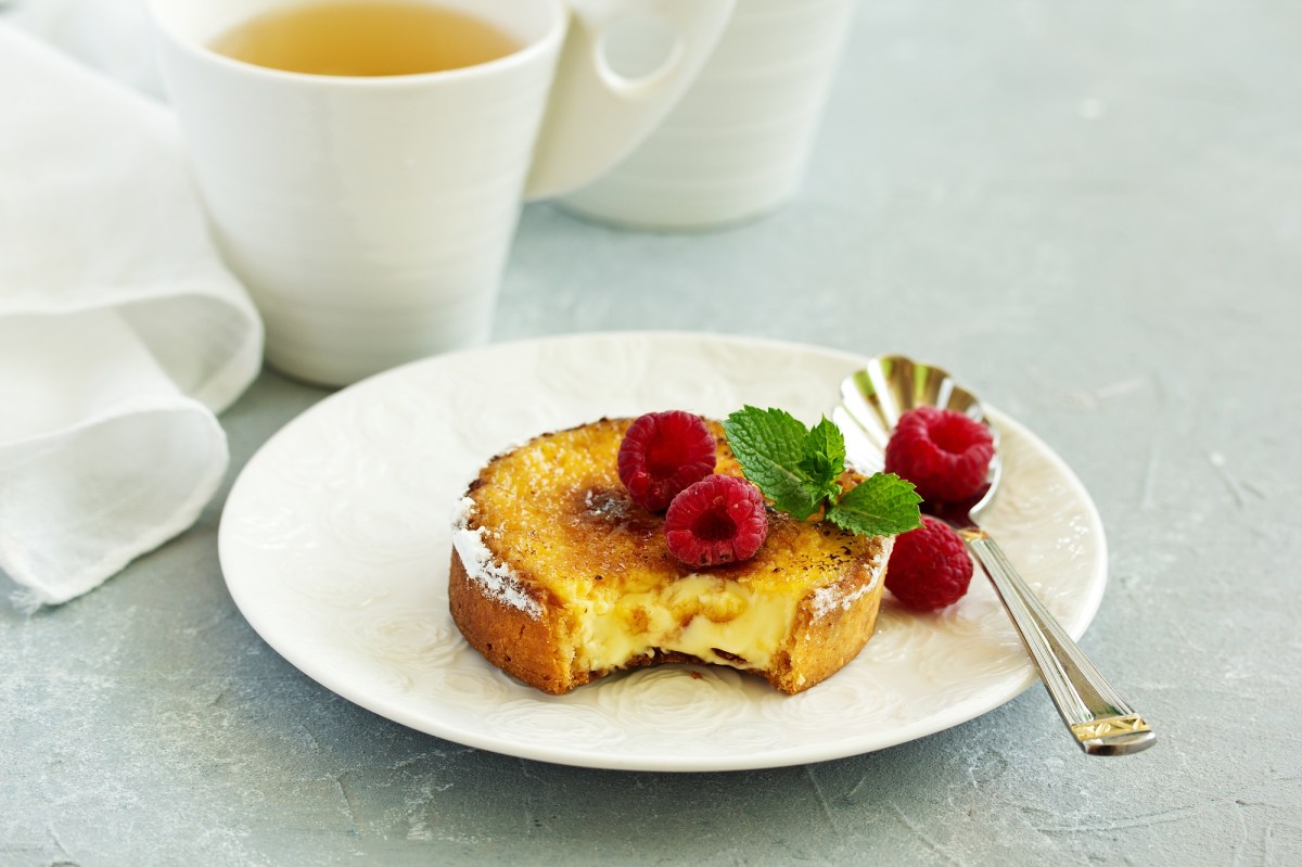 Crème-Brûlée-Kuchen, garniert mit Himbeeren und Minze, auf einem weißen Teller. Im Hintergrund steht eine Tasse Tee.