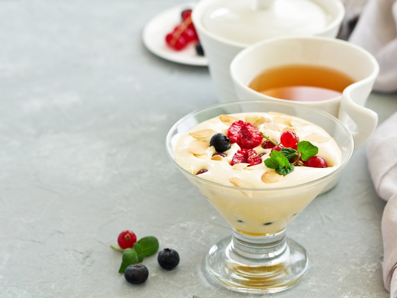 Ein Glas Eierlikör-Mousse garniert mit Beeren und Mandeln, dahinter eine Tasse Tee.