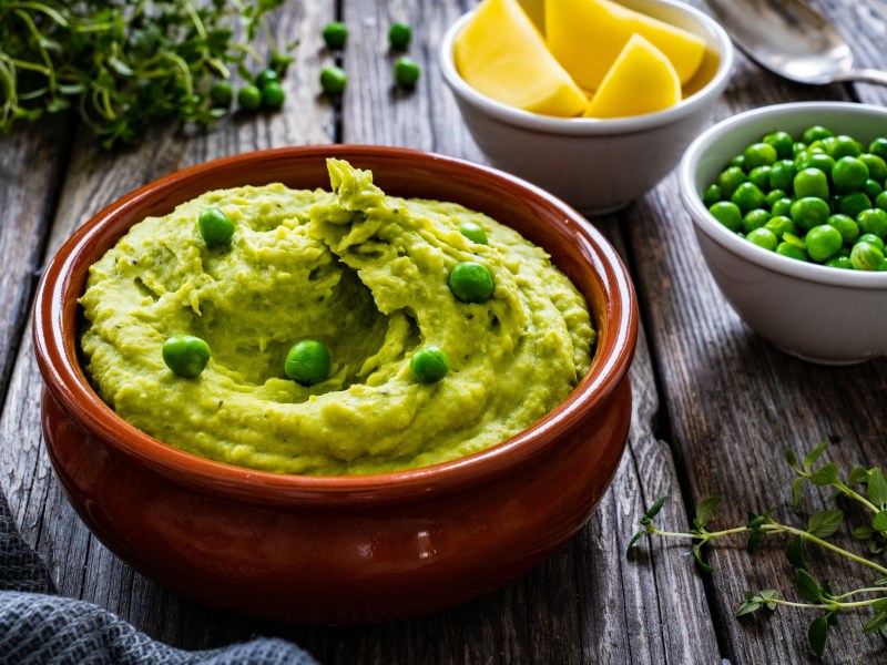 Eine braune Schale Erbsen-Guacamole, garniert mit ganzen Erbsen, daneben eine Schale Erbsen und eine Schale Zitronenspalten sowie Kräuter.