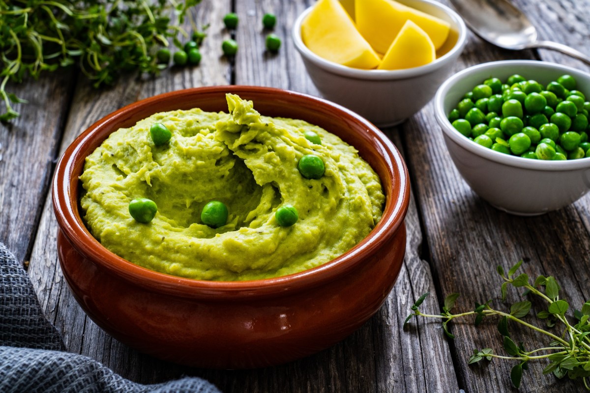 Eine braune Schale Erbsen-Guacamole, garniert mit ganzen Erbsen, daneben eine Schale Erbsen und eine Schale Zitronenspalten sowie Kräuter.