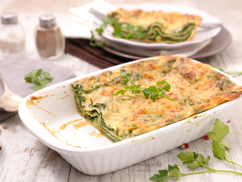 Feta-Spinat-Lasagne in einer weißen Auflaufform, zwei Stücke sind schon weg und liegen auf einem Teller im Hintergrund.