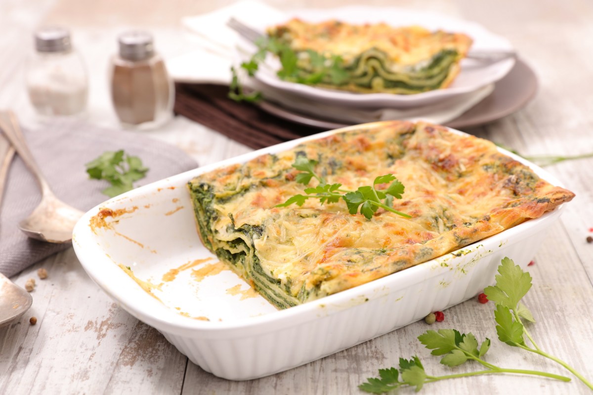 Feta-Spinat-Lasagne in einer weißen Auflaufform, zwei Stücke sind schon weg und liegen auf einem Teller im Hintergrund.