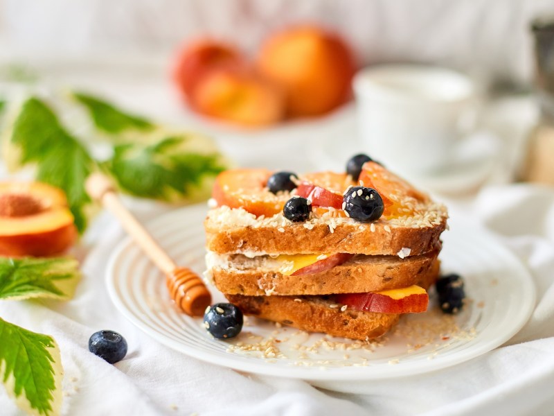 French Toasts mit gebratenen Pfirsichen, Blaubeeren und Kokosraspeln auf einem Teller gestapelt, dahinter unscharf Pfirsiche.