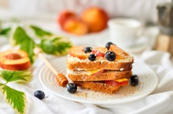 French Toasts mit gebratenen Pfirsichen, Blaubeeren und Kokosraspeln auf einem Teller gestapelt, dahinter unscharf Pfirsiche.