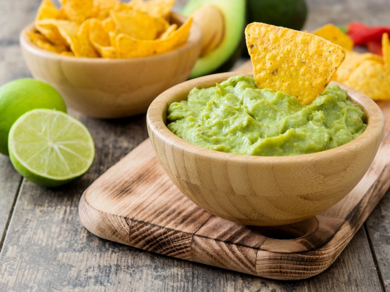 Guacamole zubereiten: eine Holzschale mit Guacamole, darin ein Tortilla-Chip, daneben eine Schale mit mehr Chips und eine halbierte Limette.